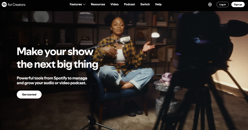 Uma pessoa est&aacute; sentada em uma cadeira azul, gravando um podcast com um microfone, cercada por prateleiras e com uma c&acirc;mera focada nela.