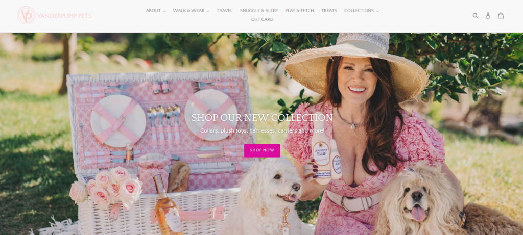 Vanderpump Pets website homepage showing a woman dressed in a pink dress sitting with two small dogs, next to a box containing picnic utensils.